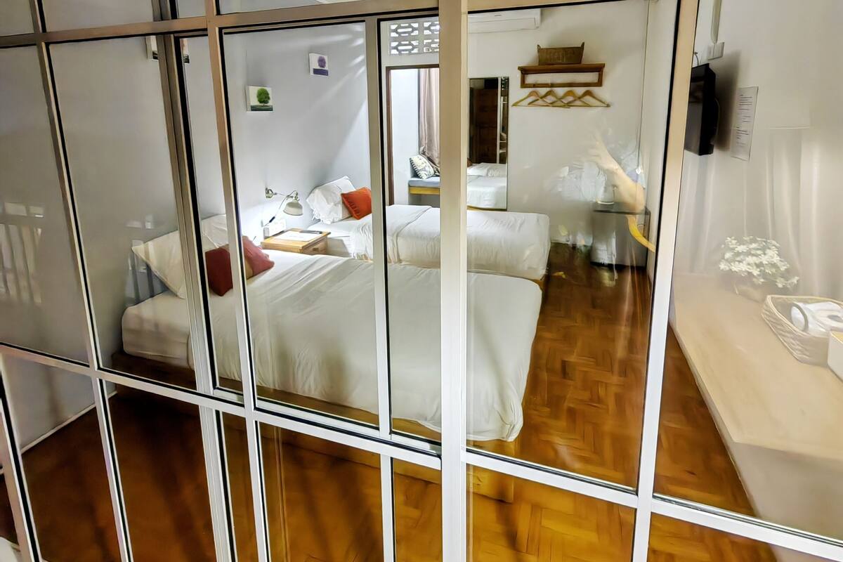 A hotel room with a wooden floor, a bed with white bedding, a bedside table, a lamp, and a small shelf with items.