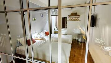 A hotel room with a wooden floor, a bed with white bedding, a bedside table, a lamp, and a small shelf with items.