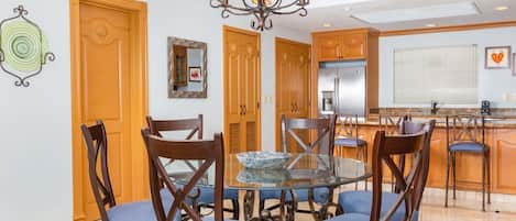 Un coin repas avec une table ronde en verre entourée de chaises en bois dotées de coussins bleus, un lustre, des portes en bois et, au fond, un espace cuisine.