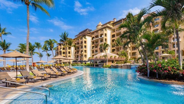 A resort pool with sun loungers, palm trees, and a multi-story building with balconies.