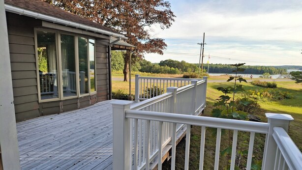 A wooden deck with white railings, a covered area with a window, and a view of a green landscape with trees and a body of water.