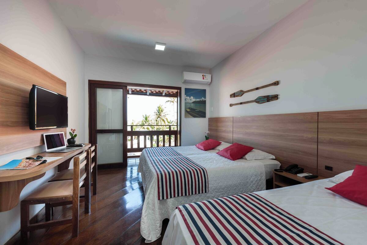 Habitación de hotel con dos camas, un escritorio con computadora, televisor y balcón con vistas a palmeras.