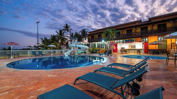 A swimming pool with lounge chairs, palm trees, and a building with a balcony in the background. - Praia Grande Hotel (Fundão)