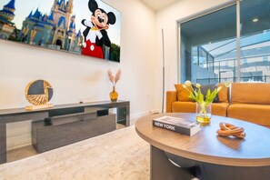 A modern living room with a large window, a flat-screen TV displaying Mickey Mouse, a round coffee table with a book titled "NEW YORK", and a sofa with a vase of flowers.