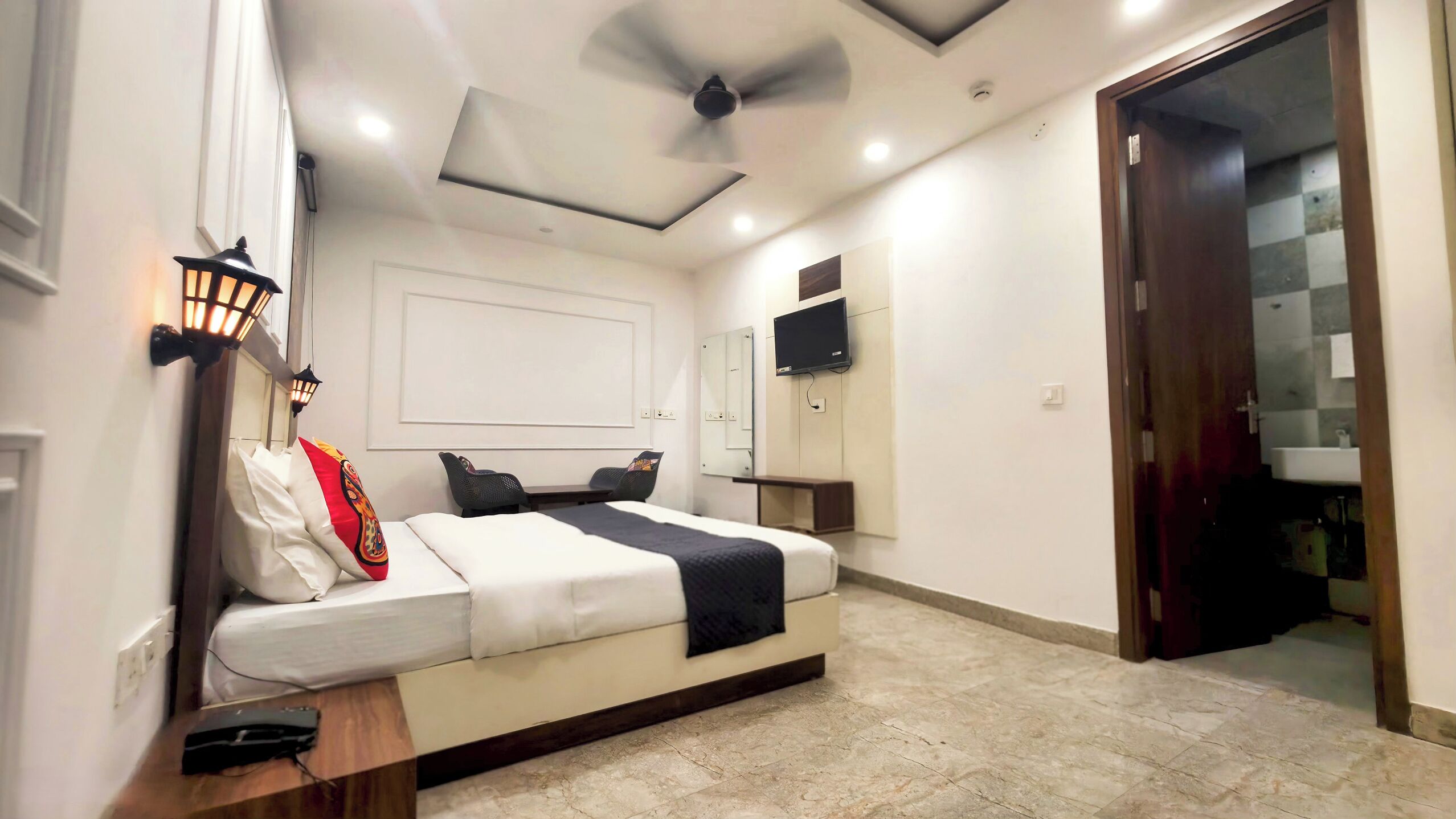 A modern hotel room with a bed, bedside table, lamp, ceiling fan, TV, and a bathroom visible through an open door.