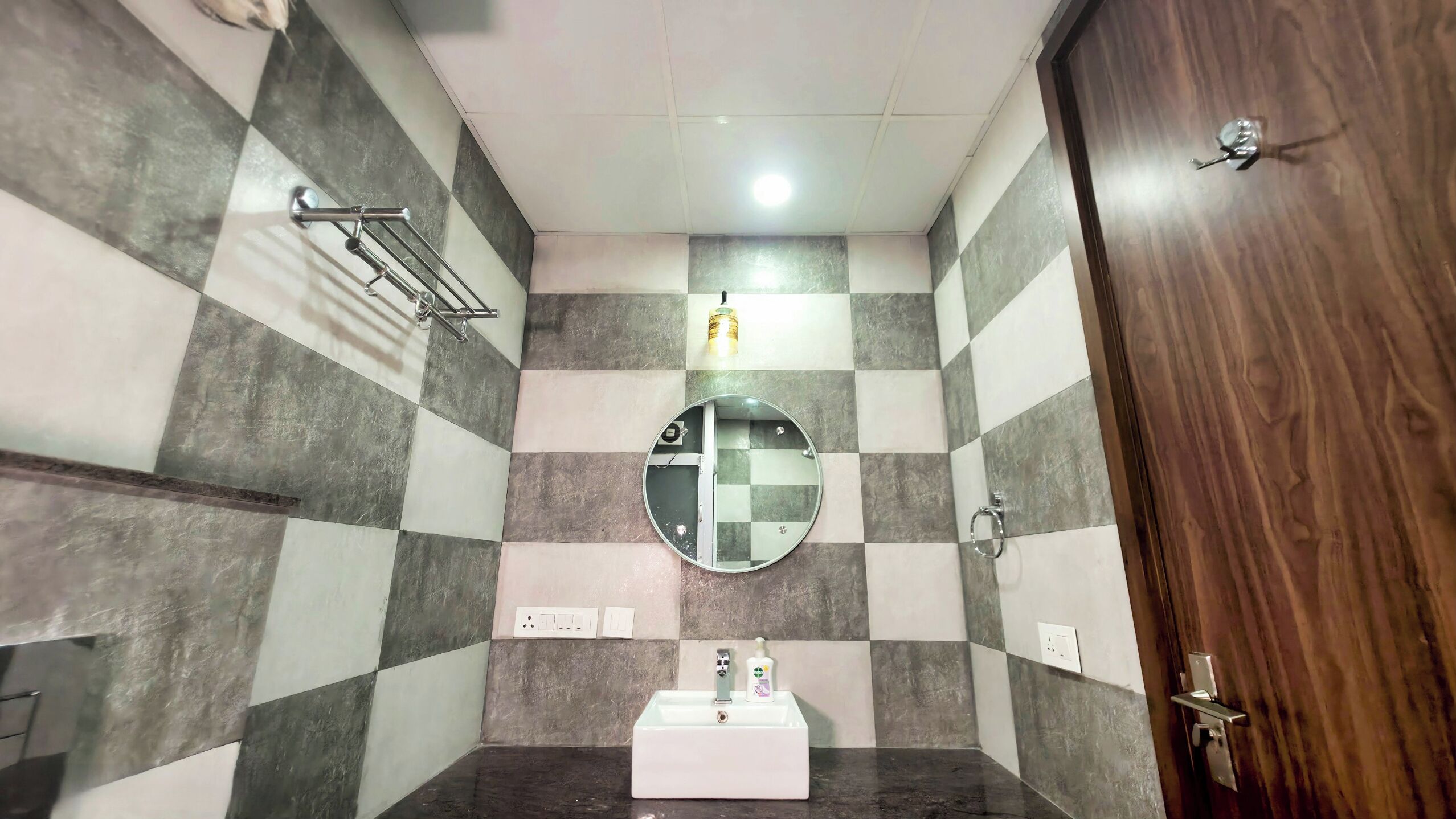 Une salle de bain avec un mur recouvert de carreaux à motifs, un miroir rond, un lavabo et une porte en bois.