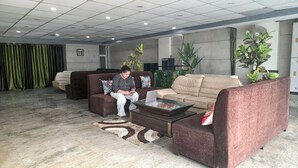 Un vestíbulo moderno con un sofá marrón, un sofá beige y un sillón marrón. Hay una mesa de centro de cristal con un ordenador portátil y un objeto decorativo. El suelo está cubierto de baldosas, y hay plantas verdes y un ventilador al fondo.