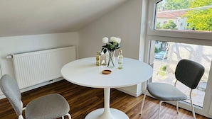 Ett litet, modernt rum med ett runt, vitt bord, två grå stolar, ett element och ett fönster med utsikt över grönska och byggnader.