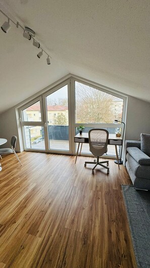 A modern room with a sloped ceiling, a desk with a chair, a sofa, and a view through large windows.