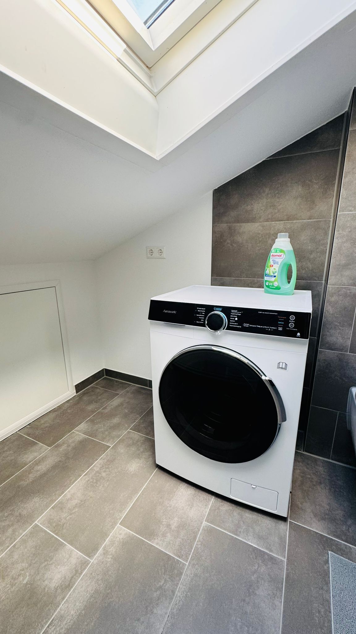 A compact, modern laundry room with a built-in washer and dryer, a skylight, and a tiled floor.