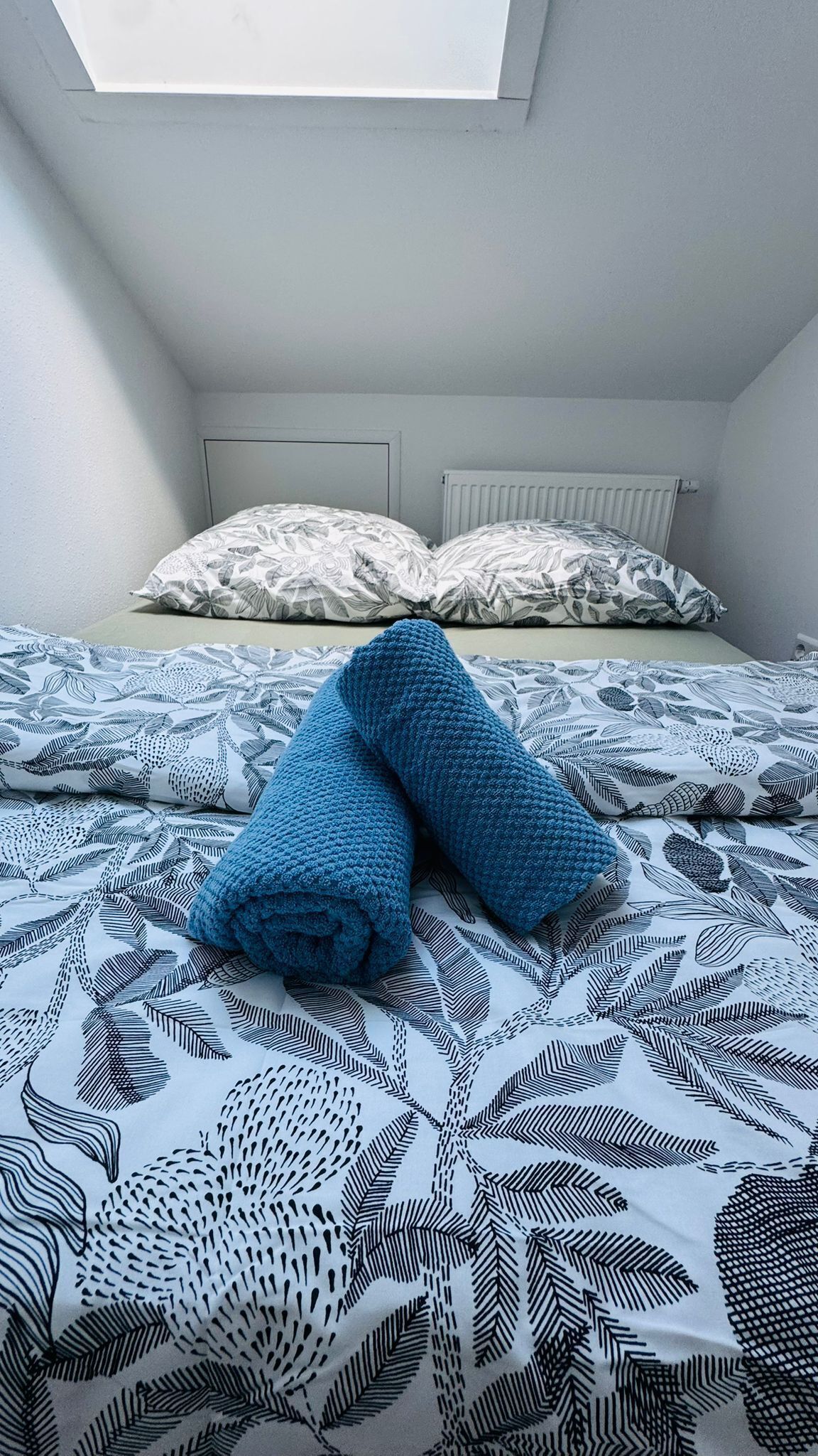 A bedroom with a bed covered in patterned bedding, a window above the bed, and a radiator on the wall.