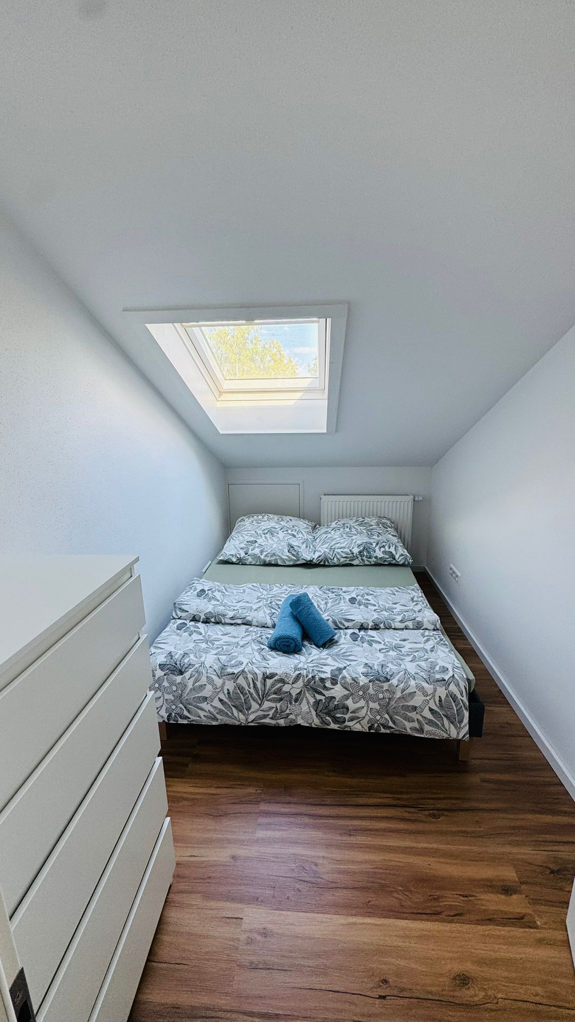 A small, well-lit room with a skylight, a bed with patterned bedding, and a wooden floor.