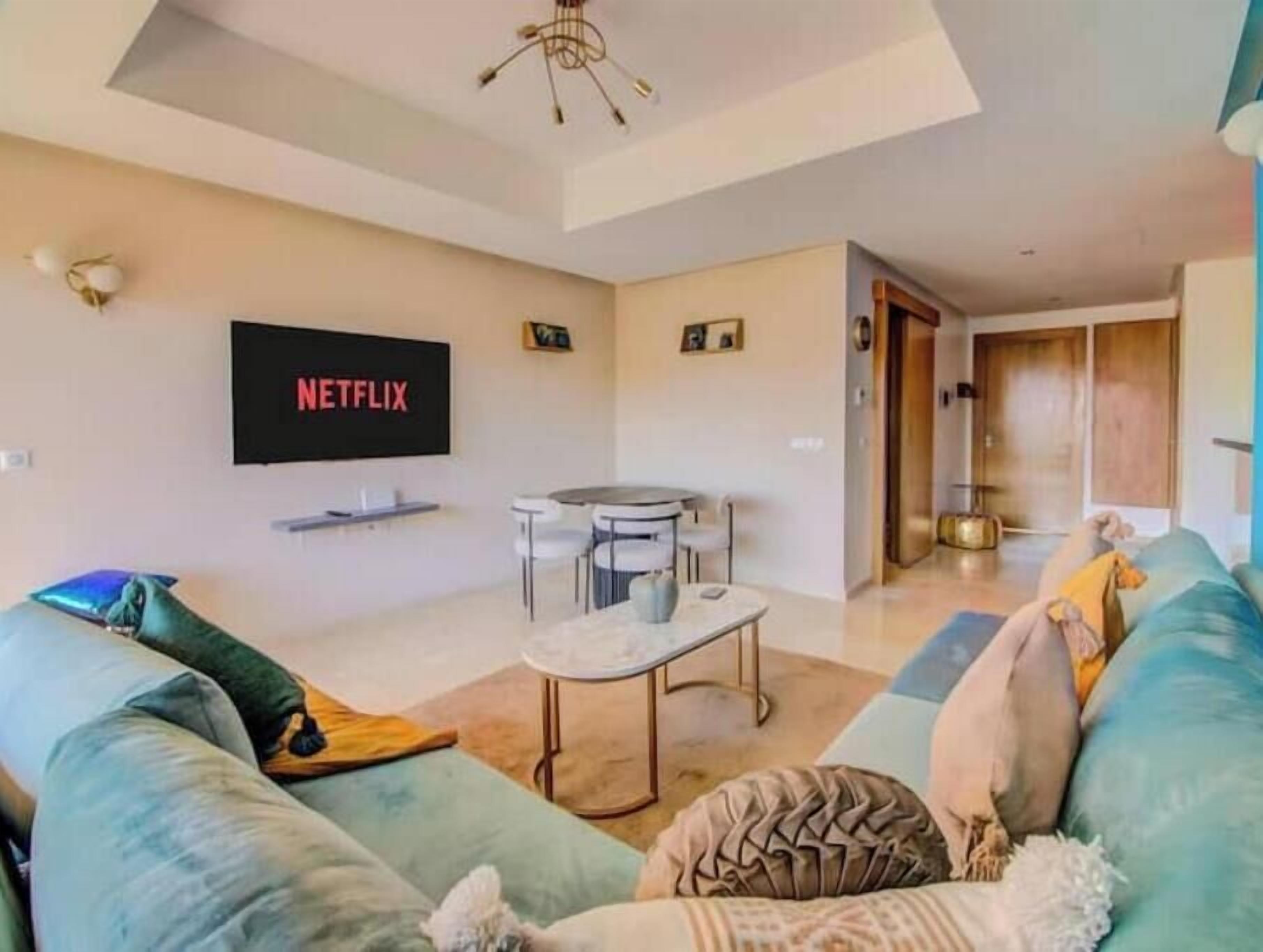 A modern living room with a flat-screen TV mounted on the wall, a round coffee table, and a sofa with cushions.