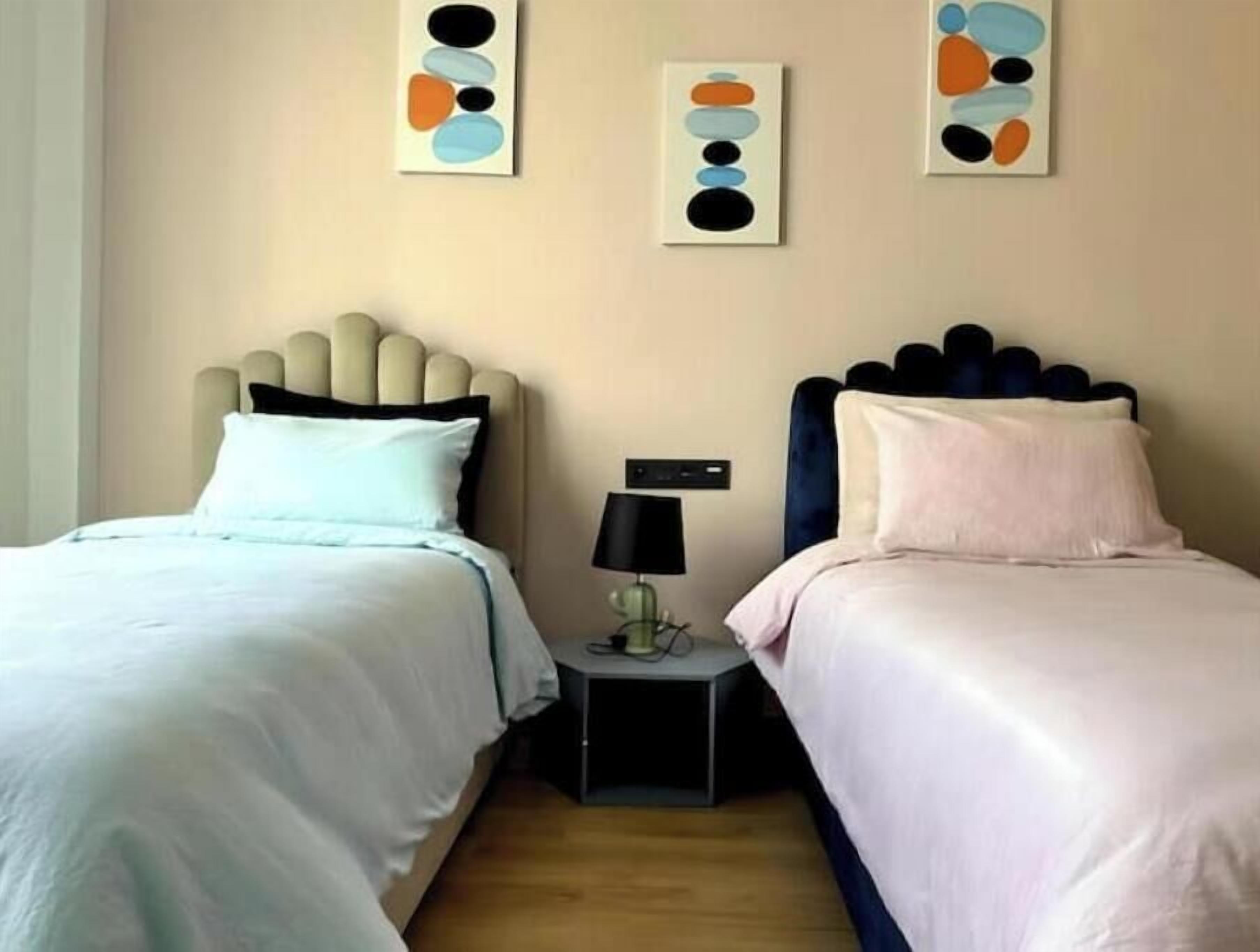 Habitació d'hotel amb dos llits, cadascun amb un disseny de capçalera diferent, una tauleta de nit amb una làmpada i una obra d'art abstracta a la paret.