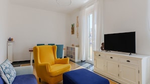 Een woonkamer met een gele fauteuil, een blauwe bank, een televisie op een witte kast en een raam met gordijnen.