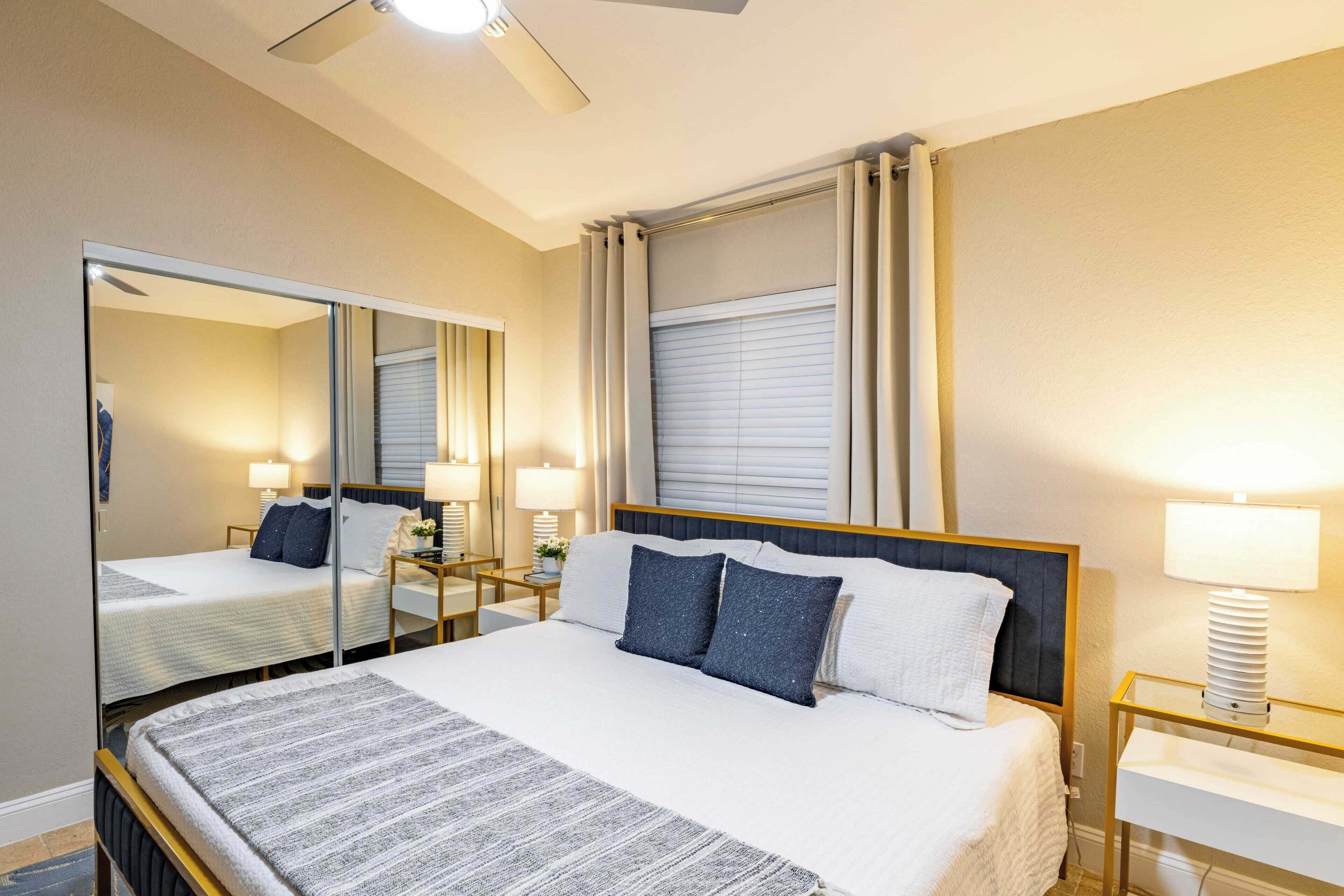 Een hotelkamer met een groot bed, een nachtkastje met een lamp, een spiegel en een raam met gordijnen.
