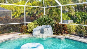 Une piscine agrémentée d’un élément rocheux, entourée de plantes tropicales et protégée par une enceinte en verre.