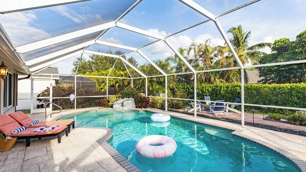 A covered outdoor swimming pool with lounge chairs, a rock feature, and a view of tropical vegetation. - Private Pool, Firepit, Game Room Mins to Beaches + Community Amenities Casa Solara by Avantstay (Naples)