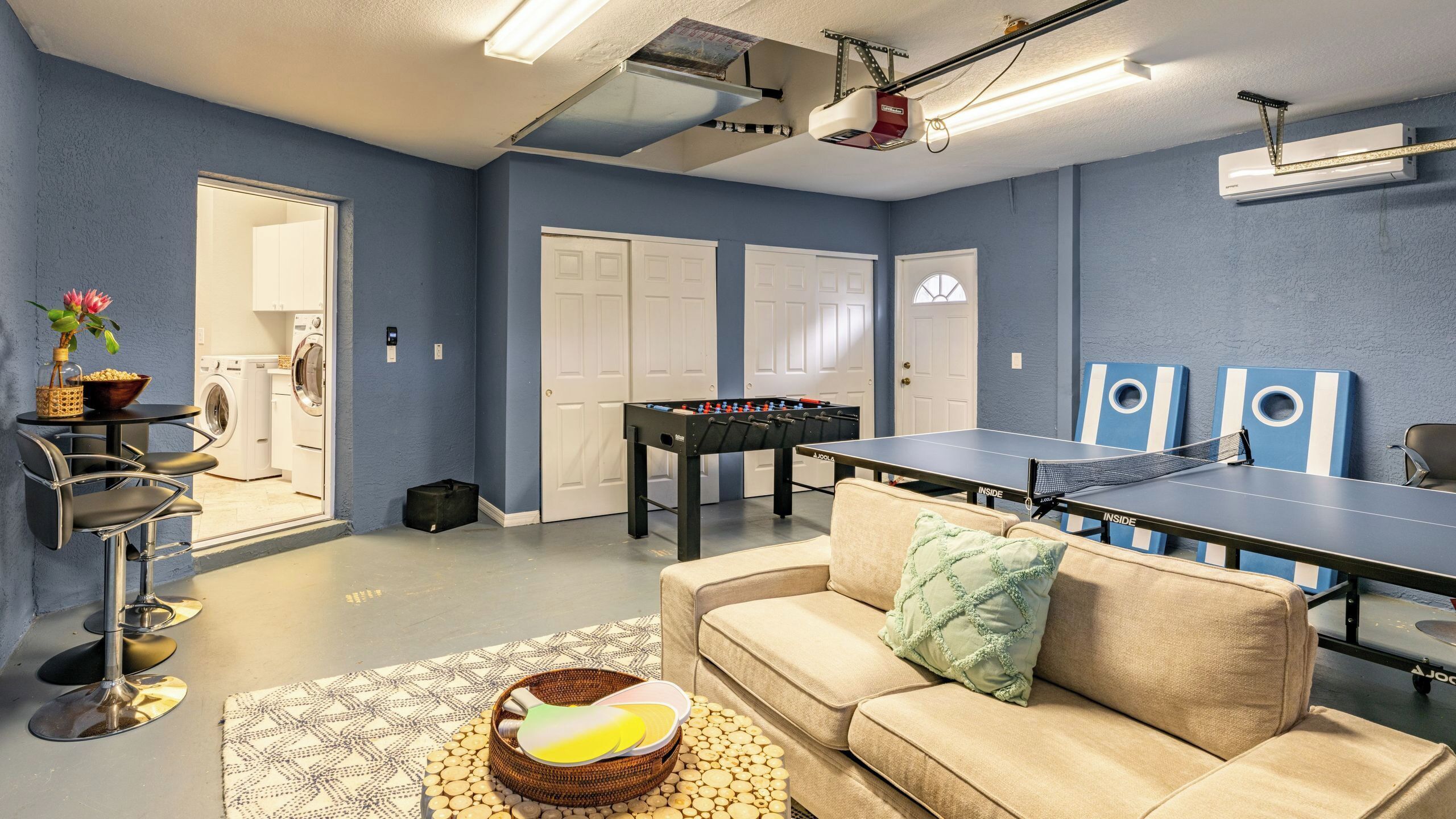 A room with a foosball table, ping pong table, sofa, and a washing machine in the background.