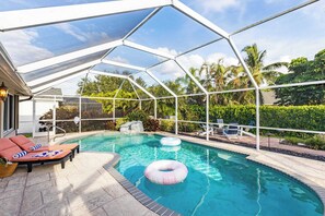 A covered swimming pool with lounge chairs and inflatable rings, surrounded by tropical vegetation. - Private Pool, Firepit, Game Room Mins to Beaches + Community Amenities Casa Solara by Avantstay (Naples)