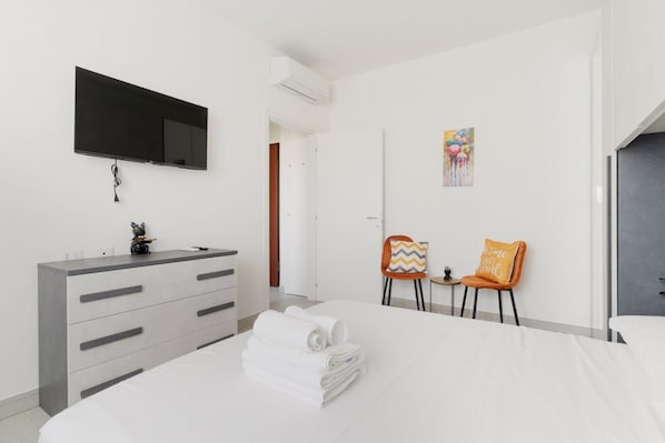 A modern hotel room with a bed, a flat-screen TV mounted on the wall, a dresser, and two chairs with cushions.