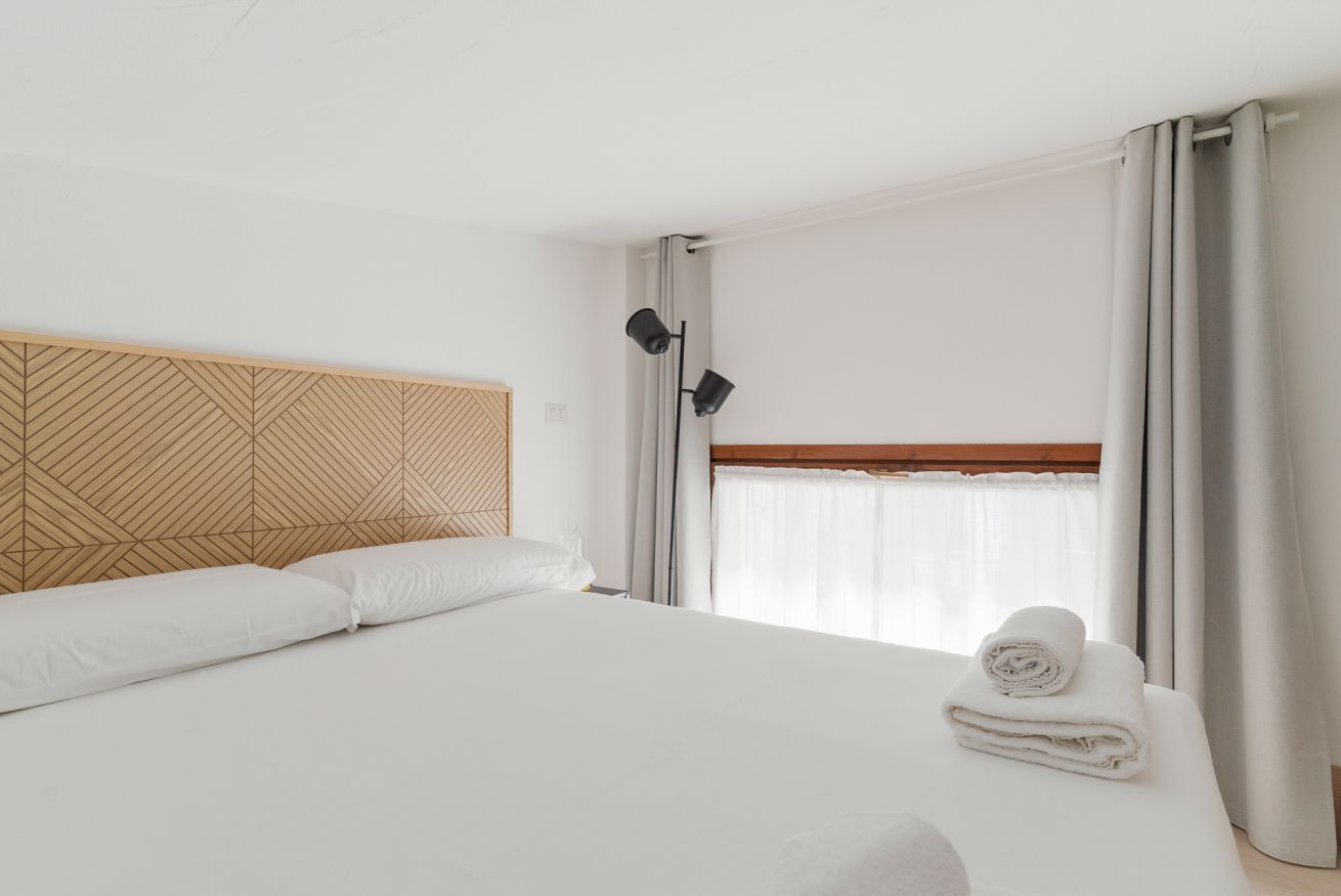 Une chambre d’hôtel avec un grand lit, une tête de lit ornée d’un motif géométrique, une fenêtre avec des rideaux et une serviette roulée posée sur le lit.