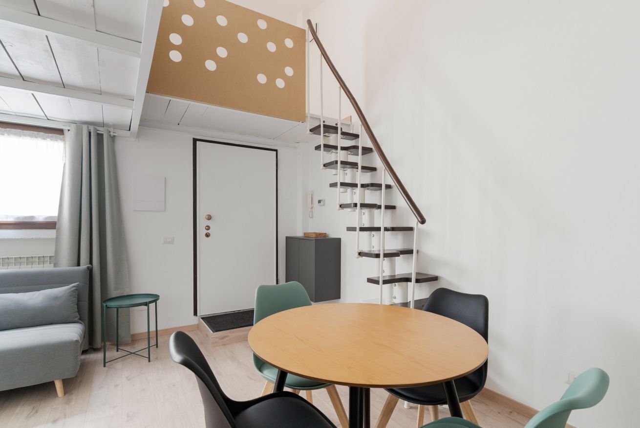 Un intérieur moderne avec une table ronde en bois, des chaises noires, un canapé gris, un petit tabouret vert, un escalier au design original et une porte blanche.