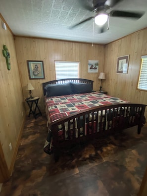 A bedroom with a wooden floor, a bed with a patterned bedspread, a ceiling fan, and two windows with blinds.