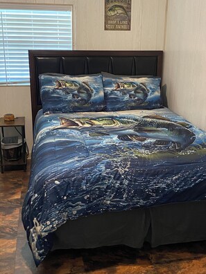Una cama con colcha y almohadas con motivos marinos azules, un cabecero oscuro y una mesita de noche de madera con una lámpara.