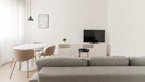 Ein modernes Wohnzimmer mit grauem Sofa, einem weißen Esstisch mit Stühlen, einem schwarzen Fernseher auf einem weißen Schrank und einer Pendelleuchte.