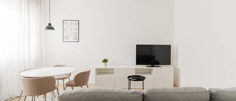Una sala de estar moderna con un sofá gris, una mesa de comedor blanca con sillas, un televisor negro sobre un mueble blanco y una lámpara colgante.