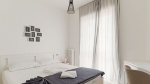 Ein ordentlich bezogenes Bett mit dunkler Decke, ein Nachttisch mit Lampe und ein Sessel.