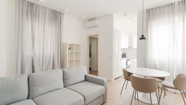 Ein modernes Wohnzimmer mit einem grauen Sofa, einem Esstisch mit Stühlen und einer Küche im Hintergrund.