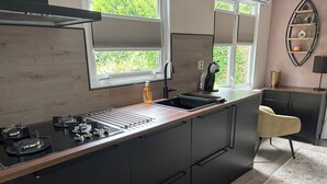 A modern kitchen with a central island, a built-in stove, and a sink. There is a chair and a decorative shelf in the background.