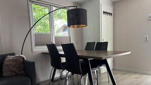 A modern living room with a dark grey sofa, a round ottoman, a wooden dining table with four chairs, a black floor lamp, and a window with blinds.