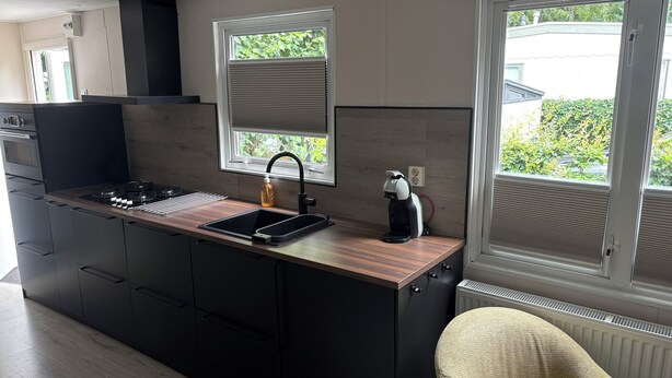 A modern kitchen with a built-in stove, a sink, and a coffee maker. There are cabinets, a window with blinds, and a radiator.