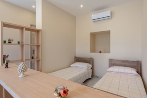 A bedroom with two beds, a wooden desk, a shelf with items, and an air conditioning unit.