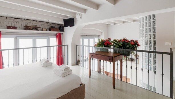 Et hotelværelse med seng, et bord med blomster, en balkon med gelænder og et fjernsyn ophængt på væggen.