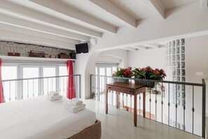 A hotel room with a bed, a table with flowers, a balcony with a railing, and a TV mounted on the wall.