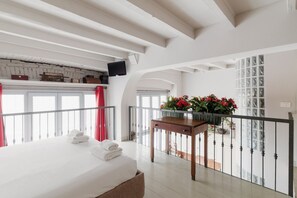 Een hotelkamer met een bed, een tafel met bloemen, een balkon met een hekwerk en een aan de muur bevestigde televisie.