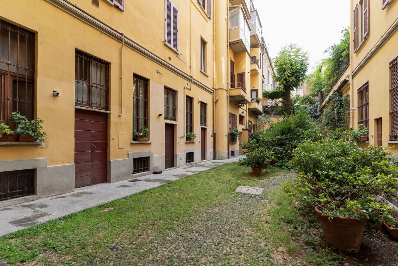 A narrow alleyway with yellow buildings, a grassy area, and potted plants.