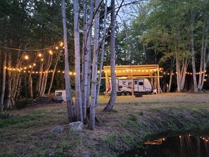 Un camping-car garé sous une structure en bois, décorée de guirlandes lumineuses, et entouré d’arbres.