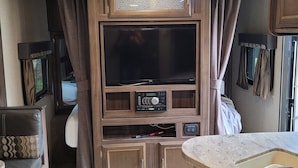 Ein kompaktes Wohnmobil-Interieur mit einem großen Flachbildfernseher, Holzschränken und einer kleinen Küchenzeile.