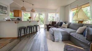 A modern living room with a grey sofa, a small kitchen area, and a large window with curtains. - Beach Bungalow 4 - 700ft to Beach - Dogs Ok! (Cannon Beach)