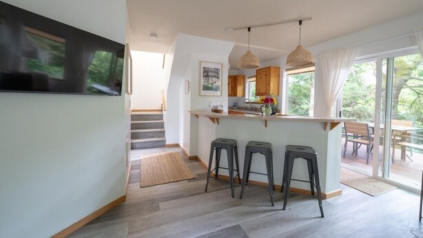 A modern kitchen with a bar area, wooden stools, and large windows offering a view of the outdoors. - Beach Bungalow 4 - 700ft to Beach - Dogs Ok! (Cannon Beach)