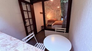 Una habitación pequeña con una mesa y sillas blancas, una puerta de madera y vistas de una cama y cortinas a través de una ventana.