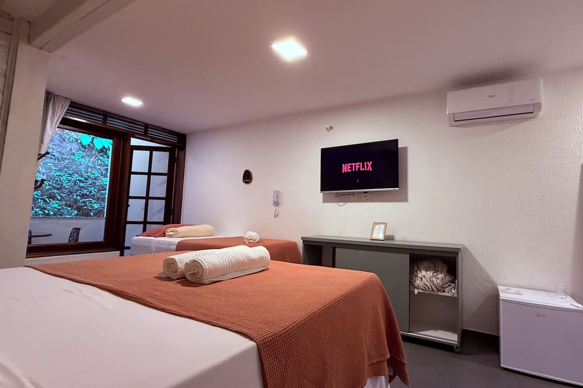 A hotel room with two beds, a TV displaying 'NETFLIX', a desk, a small fridge, and a window with a view of trees.