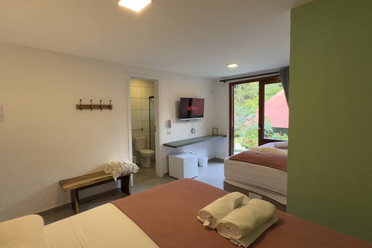 A hotel room with a bed, a bench, a desk, a television, and a window with a view of greenery.