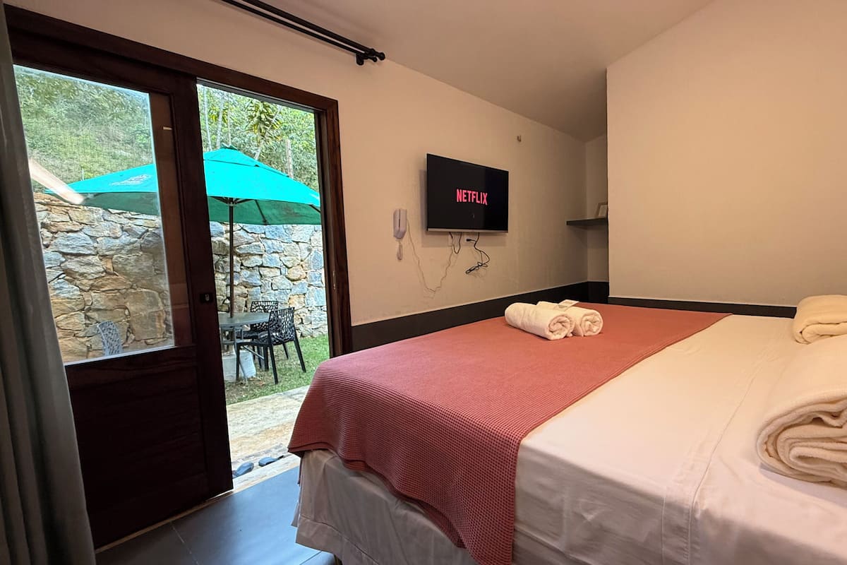 A hotel room with a bed, a TV mounted on the wall, and a view of an outdoor area with a stone wall and a green umbrella.