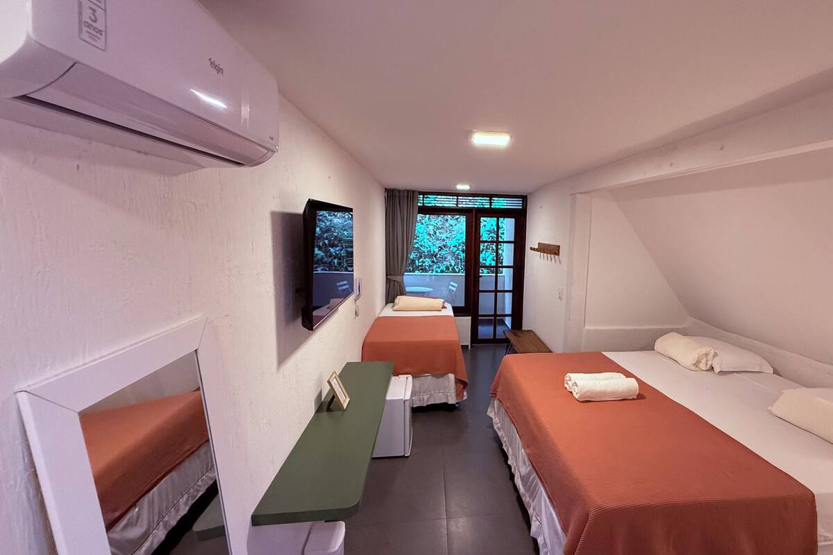 A hotel room with two beds, a desk, a television, and a balcony with a view of greenery.